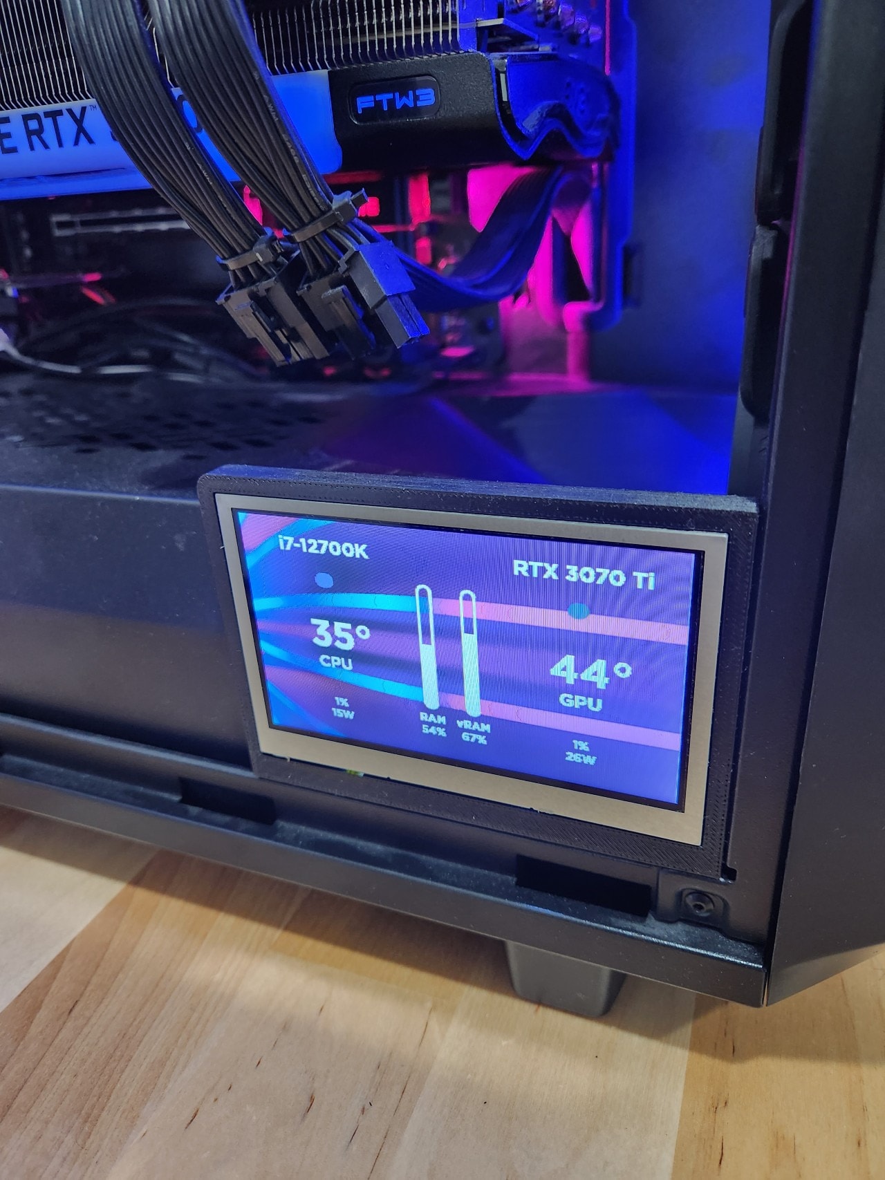 A small screen mounted low inside a dark PC case behind the glass side panel, showing CPU and GPU temperatures for an i7-12700K and an RTX 3070 Ti.